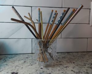 Collection of gold looking metal straws and an assortment of wooden and metal chopsticks inside a repurposed cup with dragons on it.